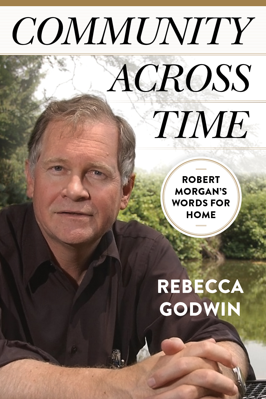 photo of the author, a white man, clean shaven, graying hair, wearing a black collared shirt with sleeves rolled up in front of a lake and trees